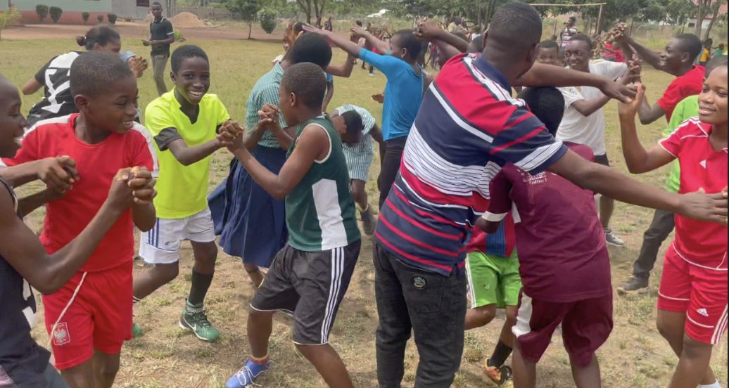 Children and adults joining raised hands into arches for the bridges game on a grass field in Ghana