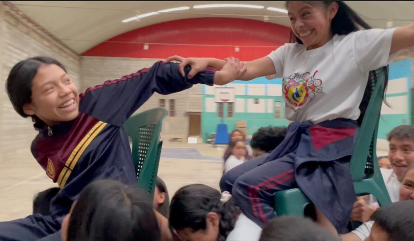 Two girls seated on chairs in a school gym, gripping each other's forearms and laughing during a chair game, classmates crowded around them