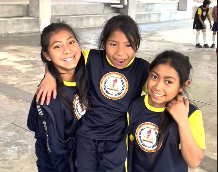 Three girls in navy and yellow school uniforms with their arms around each other, grinning at the camera