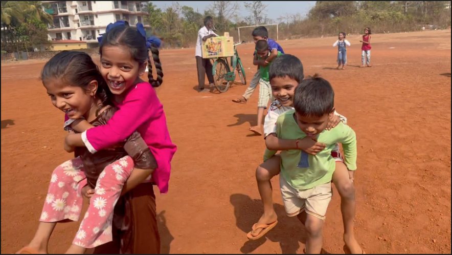 Children giving each other piggyback rides across a red dirt field in India, laughing as they run