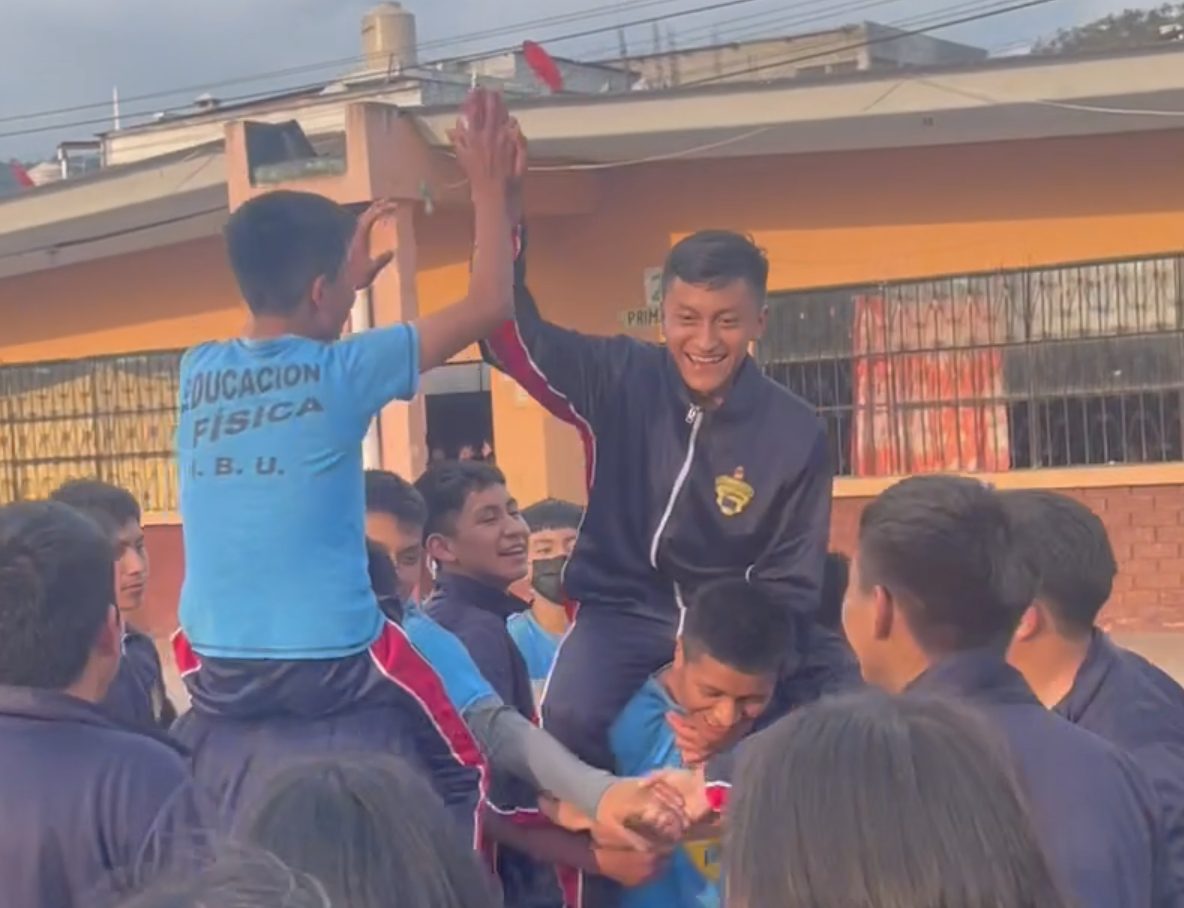 Two boys riding on classmates' shoulders and high-fiving above the crowd during a play day in Guatemala