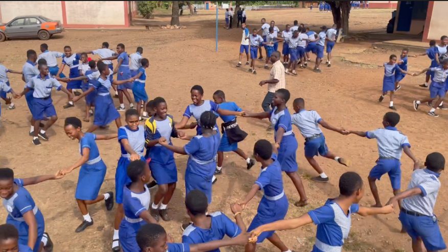 A schoolyard full of students in blue uniforms holding hands and spinning in pairs at a school in Ghana