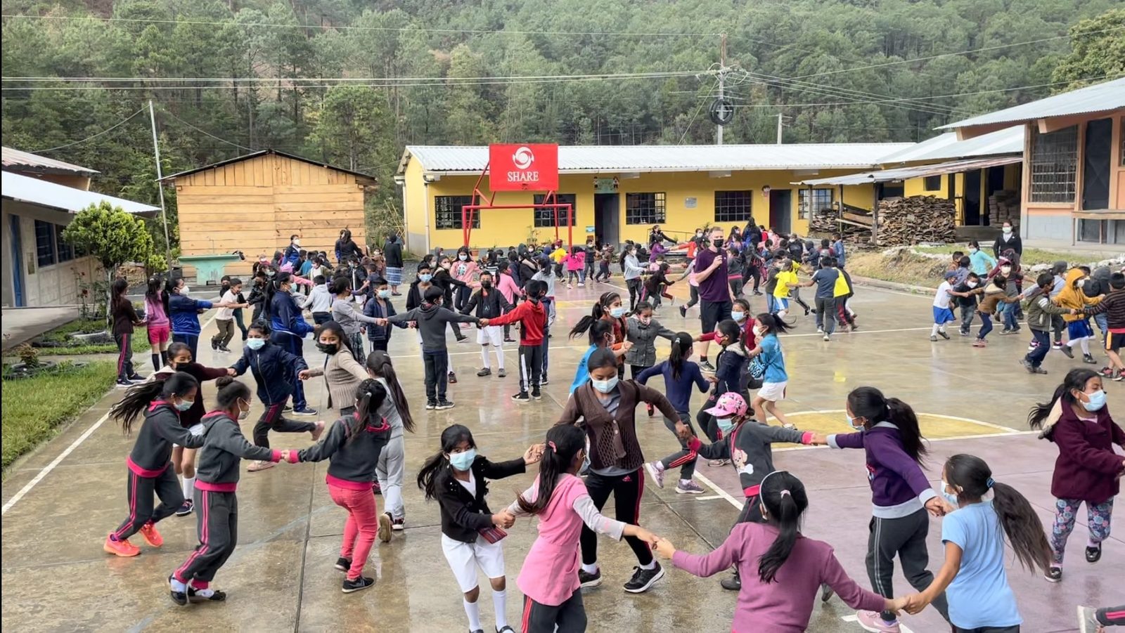 Hundreds of schoolchildren holding hands in linked circles across a concrete courtyard in Guatemala, with pine forest and mountains rising behind the school